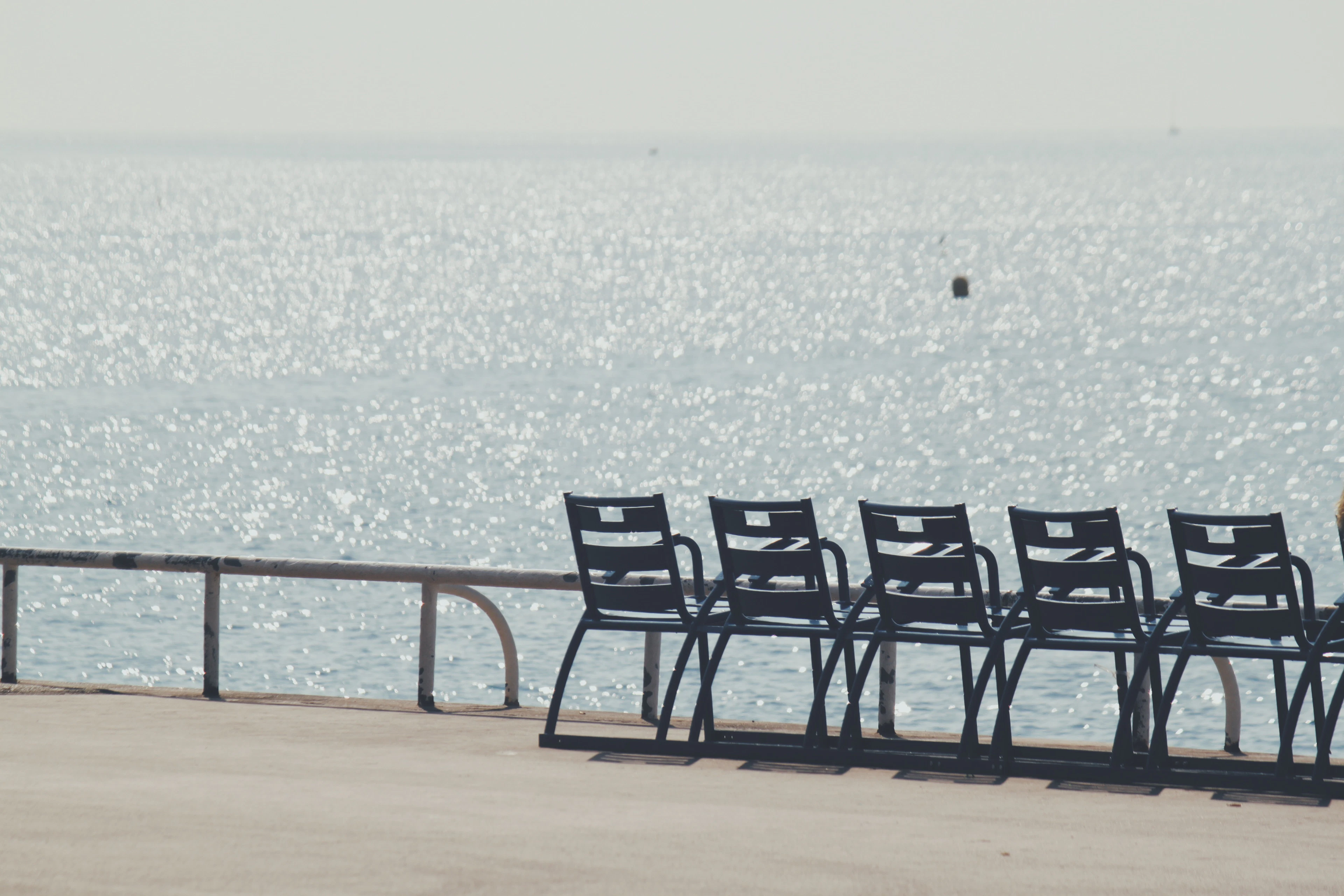 Empty blue metal chairs lined along a seafront promenade railing above a sparkling sea.
