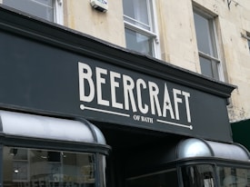 A storefront sign with the text 'BeerCraft of Bath' displayed in bold letters on a dark facade. The building is light-colored stone with two windows above the sign and a security camera mounted beside one window.