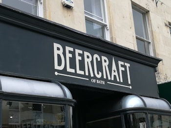 A storefront sign with the text 'BeerCraft of Bath' displayed in bold letters on a dark facade. The building is light-colored stone with two windows above the sign and a security camera mounted beside one window.