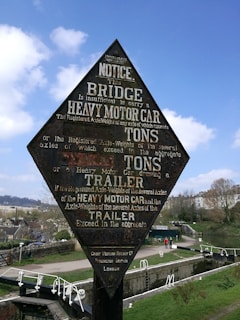 A vintage railway warning sign informs about weight limitations for vehicles on a bridge, with a scenic background of a canal lock and pathways. The sign is diamond-shaped and displays detailed text about weight restrictions. Surrounding the sign are trees, a pathway, and parts of a distant town.