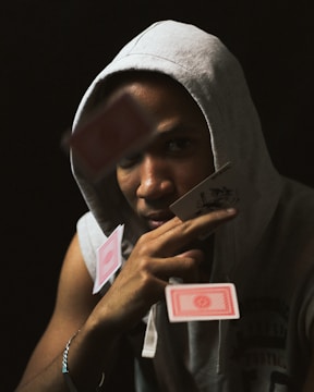A person wearing a gray hoodie is surrounded by playing cards in mid-air. The individual's face is partially obscured by a card they are holding near their eye with a contemplative expression. The background is dark, drawing focus to the person and the cards.