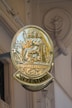 A golden sign with the emblem of a seated figure holding a staff, surrounded by detailed engravings and the words 'REPUBLIC FRANCAISE' and 'NOTAIRE'. The sign is attached to a building with ornate architectural details visible in the background.