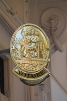 A golden sign with the emblem of a seated figure holding a staff, surrounded by detailed engravings and the words 'REPUBLIC FRANCAISE' and 'NOTAIRE'. The sign is attached to a building with ornate architectural details visible in the background.