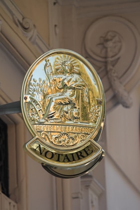 A golden sign with the emblem of a seated figure holding a staff, surrounded by detailed engravings and the words 'REPUBLIC FRANCAISE' and 'NOTAIRE'. The sign is attached to a building with ornate architectural details visible in the background.