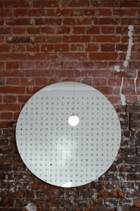 A circular mirror hanging against a brick wall with various letters and symbols printed on its surface. The mirror reflects the texture of the rough, reddish-brown bricks, adding an artistic touch to the scene.