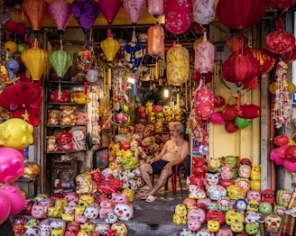 A colorful shop filled with a variety of vibrant lanterns and ceramic piggy banks in different sizes and designs. The shopkeeper, a shirtless man, is sitting on a red chair among the merchandise. The lanterns are hanging from the ceiling and are of various shapes and colors, including red, yellow, and pink. The piggy banks are arranged on the floor and shelves, featuring cartoonish faces and quirky expressions.