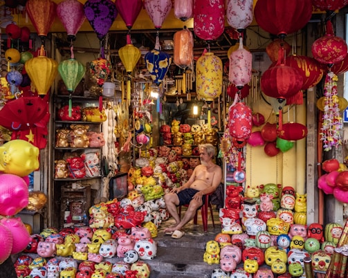 A colorful shop filled with a variety of vibrant lanterns and ceramic piggy banks in different sizes and designs. The shopkeeper, a shirtless man, is sitting on a red chair among the merchandise. The lanterns are hanging from the ceiling and are of various shapes and colors, including red, yellow, and pink. The piggy banks are arranged on the floor and shelves, featuring cartoonish faces and quirky expressions.