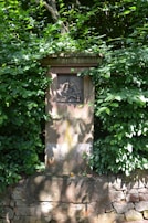 A stone monument partially covered by lush green foliage, featuring a relief carving of a person in an action-oriented pose.