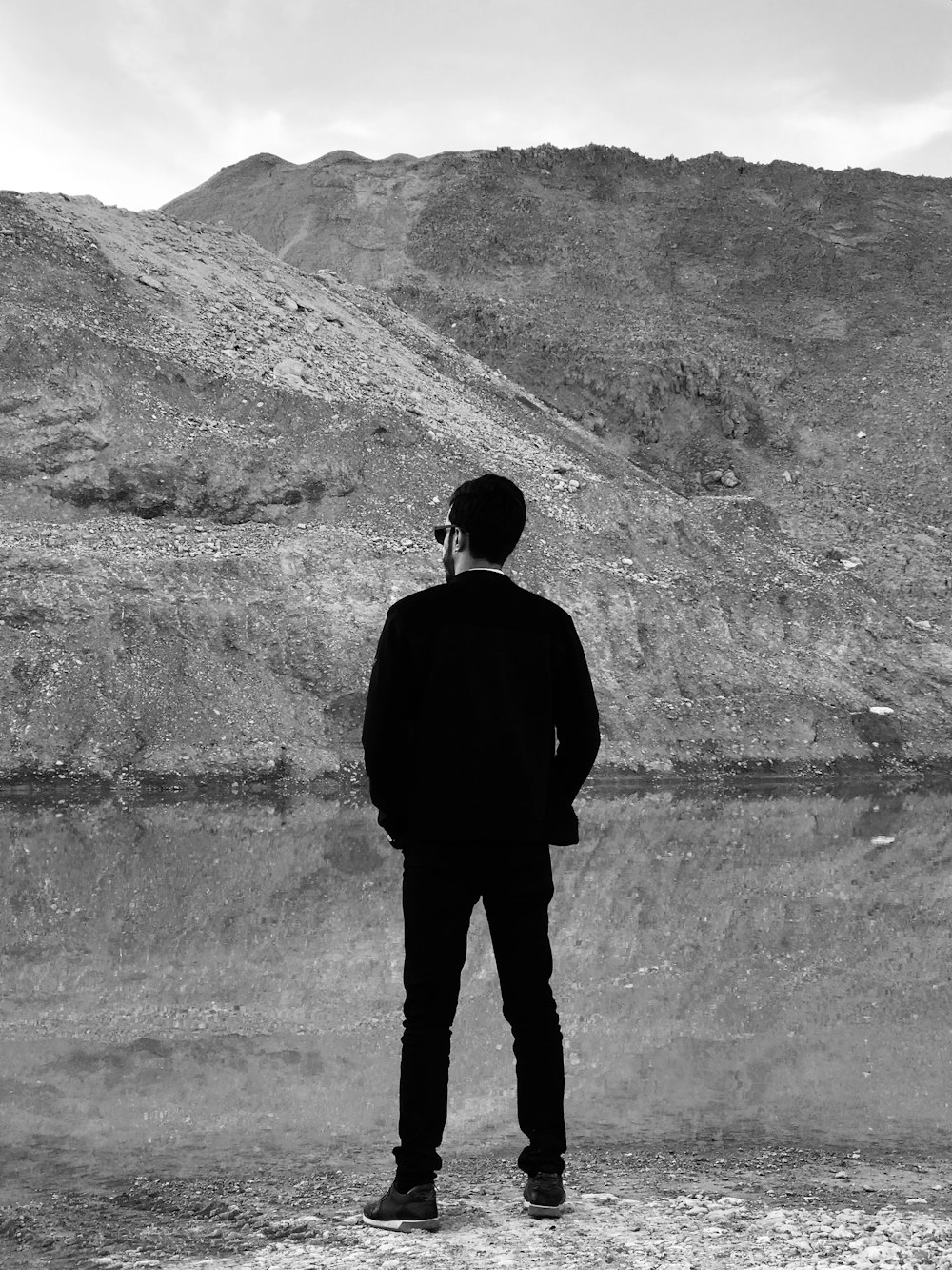 grayscale photo of man standing in front of body of water and mountain