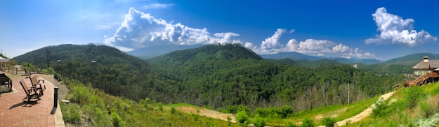 A panoramic view of lush green mountains under a bright blue sky, scattered with fluffy white clouds. There is a paved patio area with wooden chairs on the left, providing a scenic overlook. A wooden house with a sloping roof is visible on the right side, blending with the natural landscape. The landscape is expansive, showcasing rolling hills and a diverse range of vegetation.