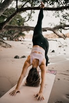 A person is practicing yoga on a sandy beach under the shade of large trees. They are performing a pose with one leg extended upward, balancing on a yoga mat. The setting is serene, with trees providing a natural backdrop and the sand suggesting a peaceful, outdoor environment.