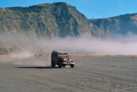 A rugged off-road vehicle drives across a vast, open sandy plain with mountains in the background and mist hovering over the landscape. The terrain appears dry and desolate, with the vehicle leaving a trail of dust behind.