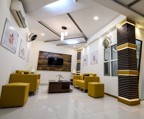 A modern and stylish waiting room featuring mustard yellow furniture including sofas and chairs. The room has a minimalist design with white walls adorned with framed art and a large clock. A flat-screen TV is mounted on a textured wooden panel. A ceiling with recessed lighting and pendant lights adds an elegant touch, and a central coffee table holds decor items.