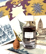 A perfume bottle labeled Boucheron Patchouli d'Angkor is surrounded by vintage prints of ancient architectural structures and Buddhist art. The background features a yellow and purple fabric with an intricate design. A small terracotta Buddha head sculpture is positioned in front of the prints.