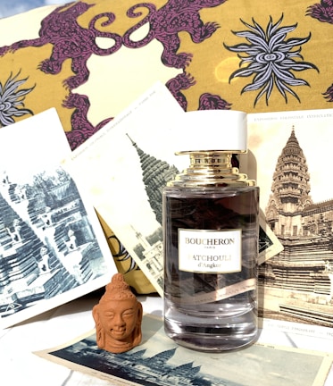 A perfume bottle labeled Boucheron Patchouli d'Angkor is surrounded by vintage prints of ancient architectural structures and Buddhist art. The background features a yellow and purple fabric with an intricate design. A small terracotta Buddha head sculpture is positioned in front of the prints.