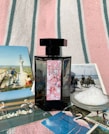 A black perfume bottle with a decorative label is placed on a striped fabric background. Surrounding the bottle are several postcards featuring scenes with flamingos, horses, and natural landscapes, as well as a small dish filled with coarse salt.