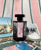 A black perfume bottle with a decorative label is placed on a striped fabric background. Surrounding the bottle are several postcards featuring scenes with flamingos, horses, and natural landscapes, as well as a small dish filled with coarse salt.