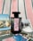 A black perfume bottle with a decorative label is placed on a striped fabric background. Surrounding the bottle are several postcards featuring scenes with flamingos, horses, and natural landscapes, as well as a small dish filled with coarse salt.