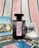 A black perfume bottle with a decorative label is placed on a striped fabric background. Surrounding the bottle are several postcards featuring scenes with flamingos, horses, and natural landscapes, as well as a small dish filled with coarse salt.