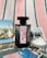 A black perfume bottle with a decorative label is placed on a striped fabric background. Surrounding the bottle are several postcards featuring scenes with flamingos, horses, and natural landscapes, as well as a small dish filled with coarse salt.