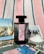 A black perfume bottle with a decorative label is placed on a striped fabric background. Surrounding the bottle are several postcards featuring scenes with flamingos, horses, and natural landscapes, as well as a small dish filled with coarse salt.