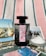 A black perfume bottle with a decorative label is placed on a striped fabric background. Surrounding the bottle are several postcards featuring scenes with flamingos, horses, and natural landscapes, as well as a small dish filled with coarse salt.