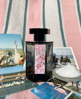 A black perfume bottle with a decorative label is placed on a striped fabric background. Surrounding the bottle are several postcards featuring scenes with flamingos, horses, and natural landscapes, as well as a small dish filled with coarse salt.