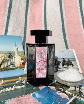 A black perfume bottle with a decorative label is placed on a striped fabric background. Surrounding the bottle are several postcards featuring scenes with flamingos, horses, and natural landscapes, as well as a small dish filled with coarse salt.