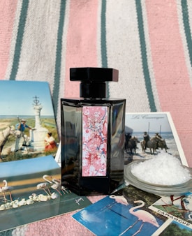 A black perfume bottle with a decorative label is placed on a striped fabric background. Surrounding the bottle are several postcards featuring scenes with flamingos, horses, and natural landscapes, as well as a small dish filled with coarse salt.