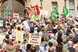 A large crowd gathers in front of a building, holding various signs and banners with messages such as 'Defend Democracy' and 'Coup des Twats.' Many people are wearing hats, and some hold green flags. The atmosphere suggests a protest or demonstration with a mix of emotions and expressions among the participants.