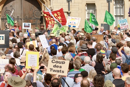 A large crowd gathers in front of a building, holding various signs and banners with messages such as 'Defend Democracy' and 'Coup des Twats.' Many people are wearing hats, and some hold green flags. The atmosphere suggests a protest or demonstration with a mix of emotions and expressions among the participants.