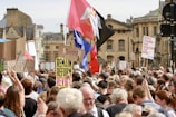 A large group of protestors gather in a city center, holding various signs and banners. The scene is bustling with people standing close together, many of whom have their backs to the camera. Signs include slogans like 'Democracy not Idiocracy' and 'Remain Reform Revolt.' Some flags are also visible among the crowd with a backdrop of historic buildings.