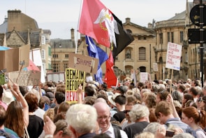A large group of protestors gather in a city center, holding various signs and banners. The scene is bustling with people standing close together, many of whom have their backs to the camera. Signs include slogans like 'Democracy not Idiocracy' and 'Remain Reform Revolt.' Some flags are also visible among the crowd with a backdrop of historic buildings.