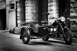 A vintage motorcycle with a sidecar is parked in an alley with rough stone walls and a brick backdrop. The setting suggests a classic, historical urban environment.