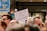 A protest or demonstration with a sign reading 'L'UNION FAIT LA FORCE' held by someone in a crowd. Other signs in the background include messages like 'DEFEND OUR DEMOCRACY'. The scene is set outdoors with a focus on activism.