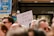 A protest or demonstration with a sign reading 'L'UNION FAIT LA FORCE' held by someone in a crowd. Other signs in the background include messages like 'DEFEND OUR DEMOCRACY'. The scene is set outdoors with a focus on activism.