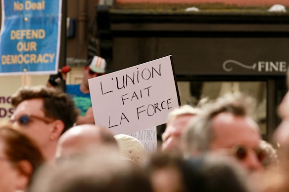 A protest or demonstration with a sign reading 'L'UNION FAIT LA FORCE' held by someone in a crowd. Other signs in the background include messages like 'DEFEND OUR DEMOCRACY'. The scene is set outdoors with a focus on activism.