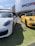 Two luxury sports cars, one white and one yellow, are parked on a grassy area outside a car dealership. The dealership has a large curved white facade with the brand's name in bold, red lettering. The cars have sleek, modern designs with shining exteriors under a clear blue sky.