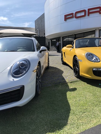 Two luxury sports cars, one white and one yellow, are parked on a grassy area outside a car dealership. The dealership has a large curved white facade with the brand's name in bold, red lettering. The cars have sleek, modern designs with shining exteriors under a clear blue sky.