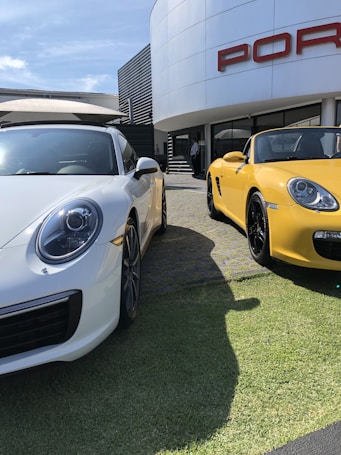 Two luxury sports cars, one white and one yellow, are parked on a grassy area outside a car dealership. The dealership has a large curved white facade with the brand's name in bold, red lettering. The cars have sleek, modern designs with shining exteriors under a clear blue sky.