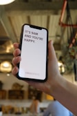 A hand holds a smartphone displaying a motivational message that reads 'It's 9AM and you're happy.' The screen background is white with the text in bold red letters. In the background, a blurred indoor setting with warm lighting and decor elements can be seen.