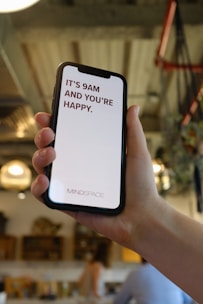 A hand holds a smartphone displaying a motivational message that reads 'It's 9AM and you're happy.' The screen background is white with the text in bold red letters. In the background, a blurred indoor setting with warm lighting and decor elements can be seen.