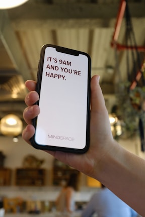A hand holds a smartphone displaying a motivational message that reads 'It's 9AM and you're happy.' The screen background is white with the text in bold red letters. In the background, a blurred indoor setting with warm lighting and decor elements can be seen.