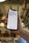A hand holds a smartphone displaying a motivational message that reads 'It's 9AM and you're happy.' The screen background is white with the text in bold red letters. In the background, a blurred indoor setting with warm lighting and decor elements can be seen.