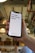 A hand holds a smartphone displaying a motivational message that reads 'It's 9AM and you're happy.' The screen background is white with the text in bold red letters. In the background, a blurred indoor setting with warm lighting and decor elements can be seen.