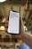 A hand holds a smartphone displaying a motivational message that reads 'It's 9AM and you're happy.' The screen background is white with the text in bold red letters. In the background, a blurred indoor setting with warm lighting and decor elements can be seen.