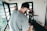 A person with a beard and cap is handling a professional camera, filming in an indoor space with bright natural lighting. In the background, two individuals stand near a drum set, suggesting a music or video production setting. Large glass windows allow natural light to flood the room.