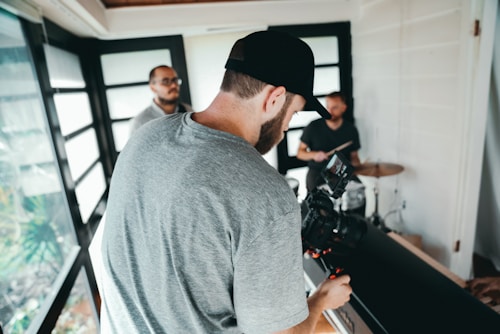 A person with a beard and cap is handling a professional camera, filming in an indoor space with bright natural lighting. In the background, two individuals stand near a drum set, suggesting a music or video production setting. Large glass windows allow natural light to flood the room.