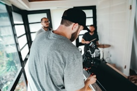 A person with a beard and cap is handling a professional camera, filming in an indoor space with bright natural lighting. In the background, two individuals stand near a drum set, suggesting a music or video production setting. Large glass windows allow natural light to flood the room.