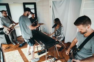A group of five people are in a small room, engaged in a musical performance. One person is seated at a keyboard, singing into a microphone, while others are playing guitars and a drum set. Recording equipment is set up in front of them, capturing the performance.